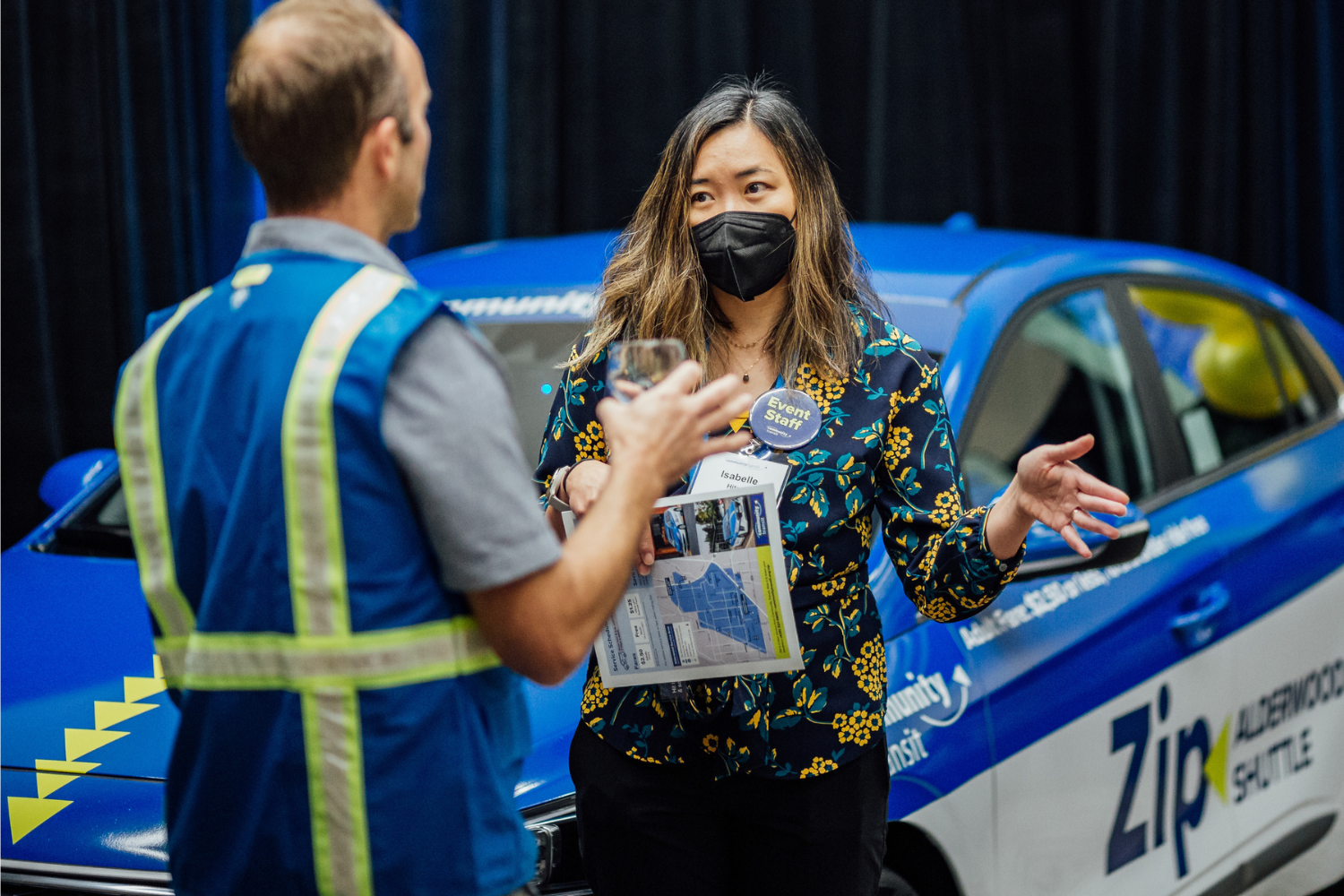 A man and a woman stand having a conversation near a Community Transit Zip Alderwood Shuttle car at an October 2022 launch event.