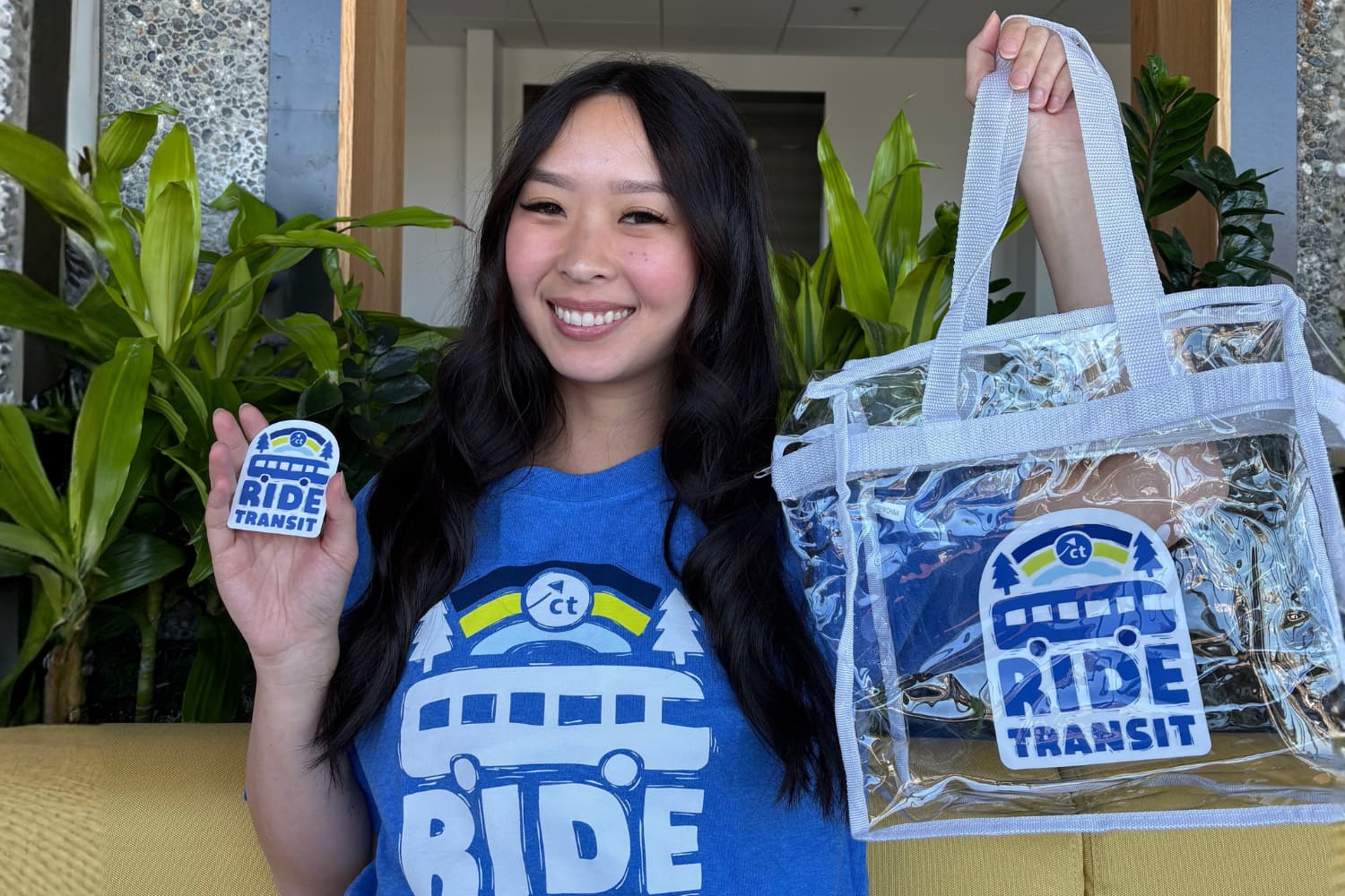 A Asian woman with long black hair wears a Ride Transit Month tshirt and holds matching merchandise that can be won as prizes