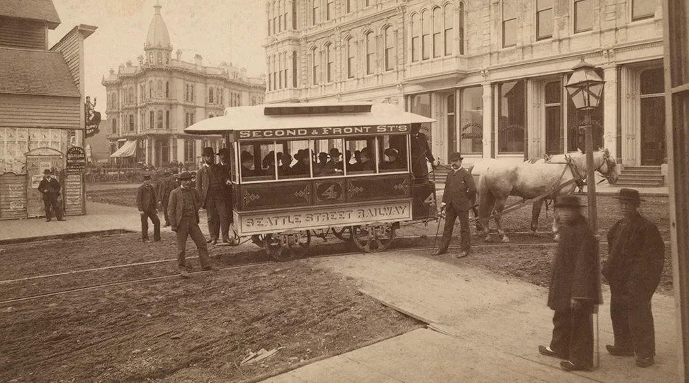 Black-and-white photo from September 1884 showing the Seattle Street Railway’s first horse-drawn streetcar at Mill Street (now Yesler Way) and Second Avenue. Mayor John Leary and dignitaries are seated inside the car on opening day. Tracks run through a dirt road, and two pedestrians observe from a wooden sidewalk on the right. This marked the beginning of streetcar service in Washington Territory.