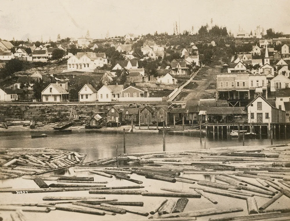 Black-and-white 1880 photo taken from Elliott Bay, looking up Columbia Street from Yesler’s Wharf. A scattering of modest, tide-worn shacks—likely built from salvaged materials—line the unpaved street that climbs the hill. Logs float in the foreground near the shore, awaiting milling. The early urban landscape predates the fire of 1889 and the redevelopment that transformed the area into a transportation hub. 