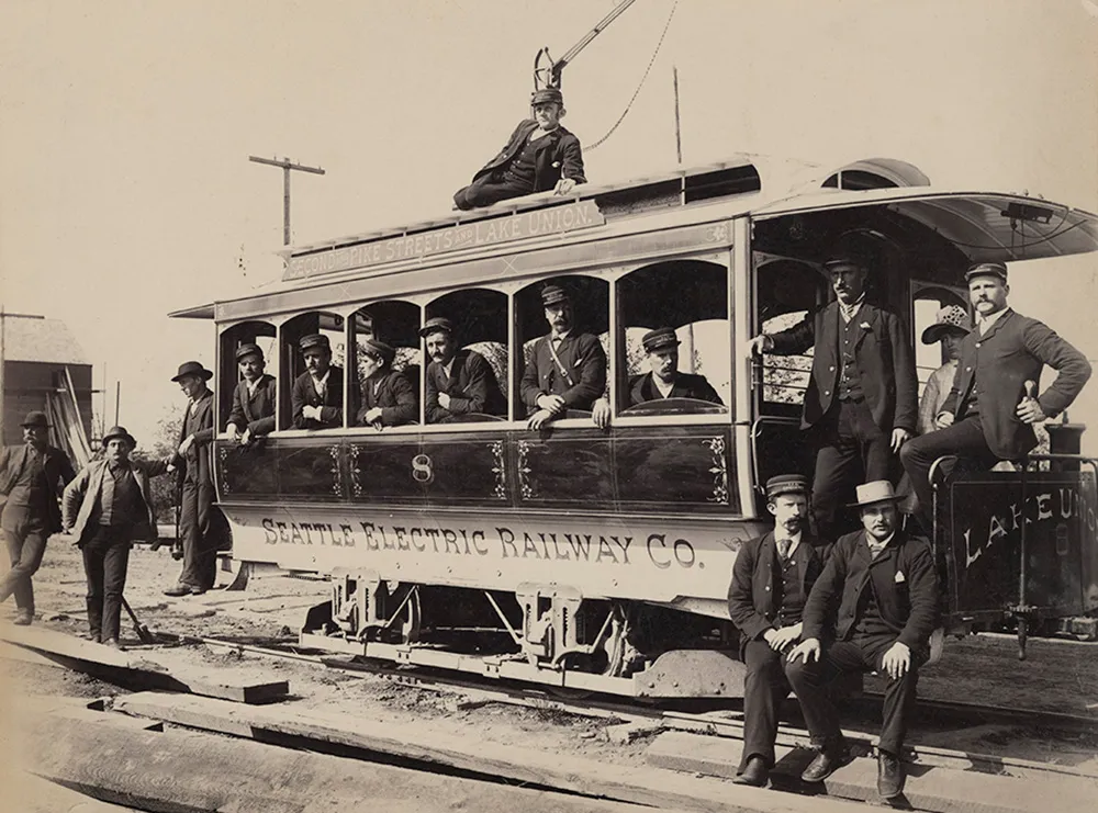 Black-and-white photo from around 1890 showing motormen from the Seattle Electric Railway and Power Company standing beside and aboard Streetcar #8. Taken near the start of electric streetcar service to Lake Union, the image reflects early transit labor conditions and urban development. Handwritten notes mention W.X. Wright and the Eastlake Line.