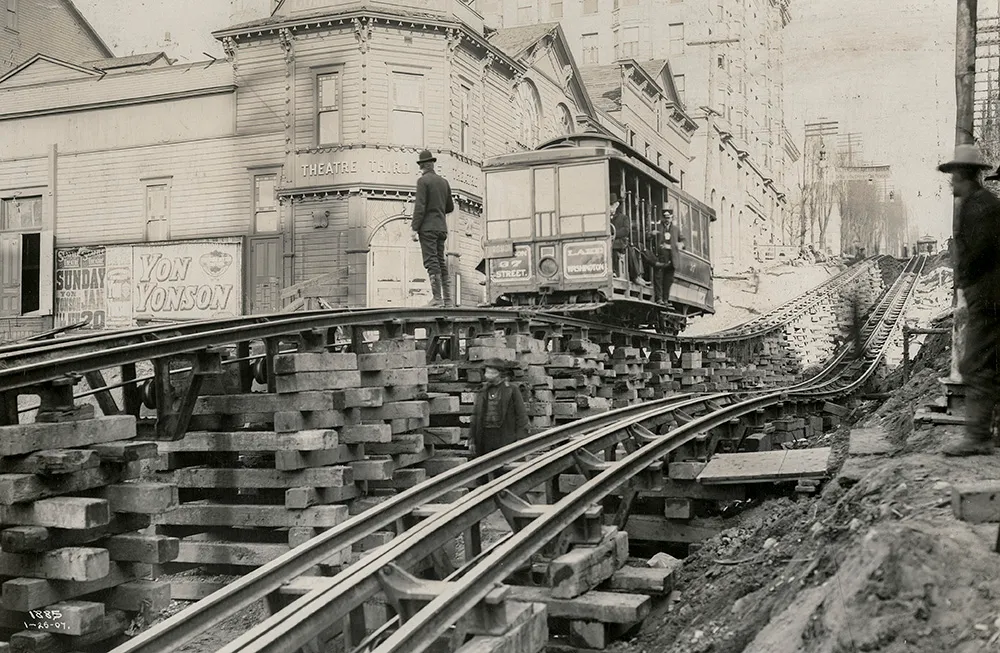 Black-and-white photo from 1907 showing a Madison Street Cable Railway car elevated on timber cribwork during the Third Avenue regrade in downtown Seattle. A conductor stands beside the car, and a second man waits near the tracks. In the background, the Seattle Theatre advertises a Yon Yonson performance, and the Lincoln Hotel is visible uphill at Fourth and Madison.