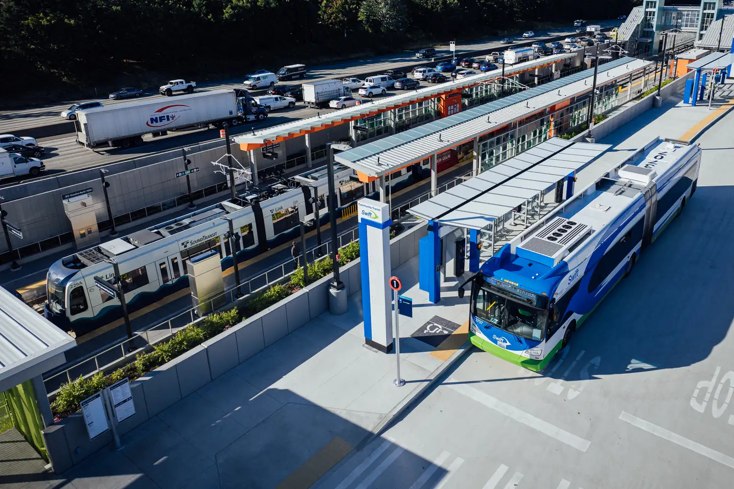 a Swift bus stopped at a light rail station with a Link light rail train on the nearby tracks